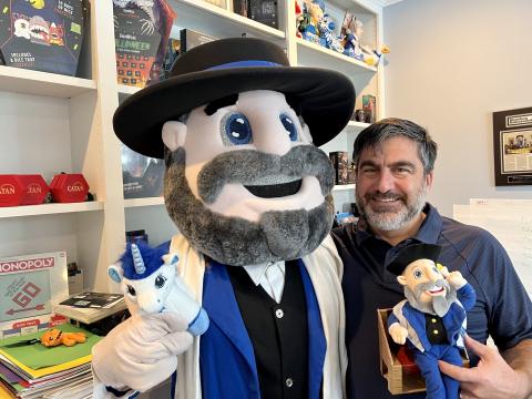 Neal Hoffman stands with the Moshe the Mensch mascot in his home office. He holds a Mensch on a Bench in his hand.