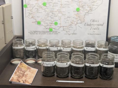 The temporary display at the Underground Railroad Museum displays 14 jars, each filled with the dirt from a lynching site in Ohio. The jars bear the names of the Black Ohioans who were lynched at those locations. The green dots on the map mark where each lynching happened.